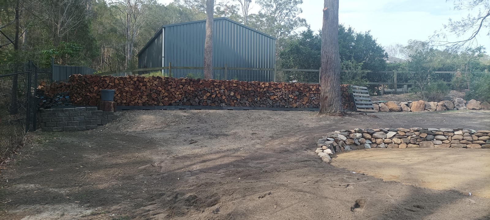 A bush block cleared back to open ground, with split firewood stacked along the fence and a stone edge